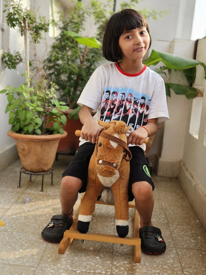 A young girl rides a toy horse on a balcony surrounded by potted plants, exuding joy and playfulness.